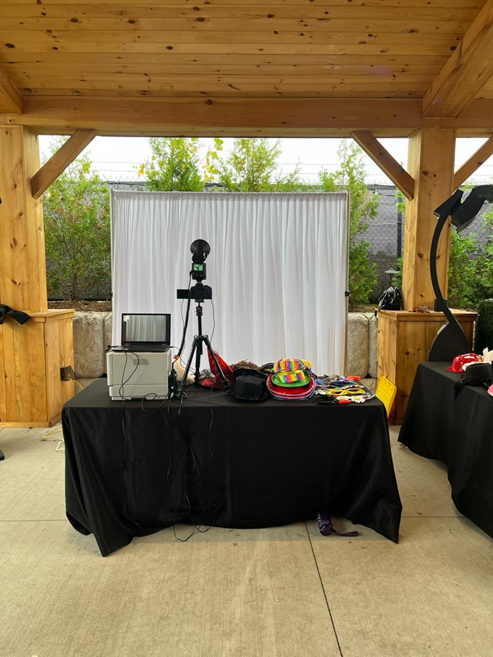 WithNoir open-air DSLR booth set up under a wooden pavilion — white drape backdrop, camera on tripod, and props table, ready before guests arrive