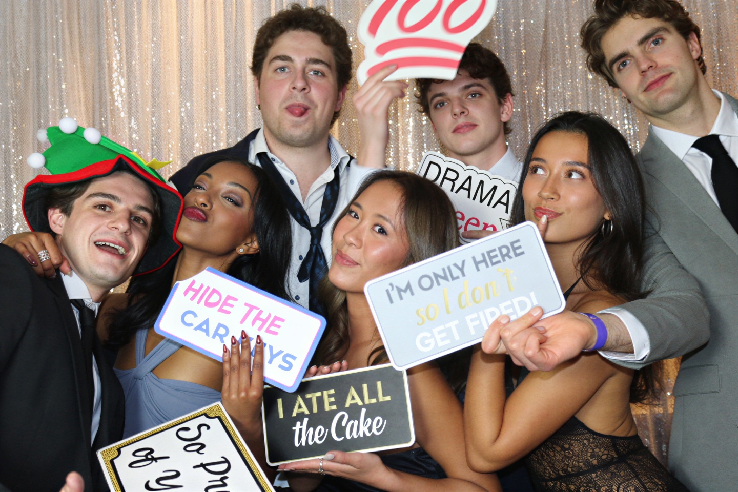 A group of friends laughing with props and signs in front of a champagne sequin backdrop